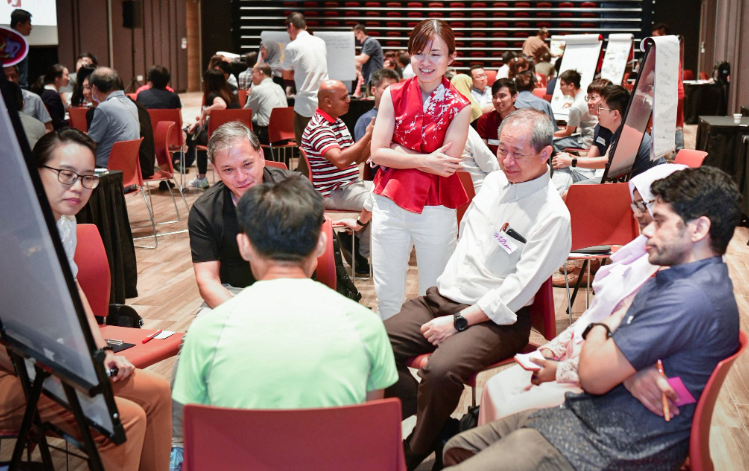 A group of people are seated in a circle during a workshop in a room with red chairs and presentation boards.